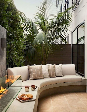 A cozy outdoor patio with a curved built-in sofa, neutral cushions, a small fire pit, and tall tropical plants against a modern wall.