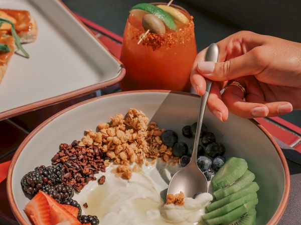 A bowl with yogurt, granola, strawberries, blueberries, kiwi, banana, and a drink beside it. A hand holds a spoon over the bowl.