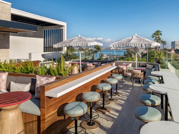The image shows a rooftop terrace with tables, chairs, and umbrellas, overlooking a scenic view of the ocean and distant buildings.