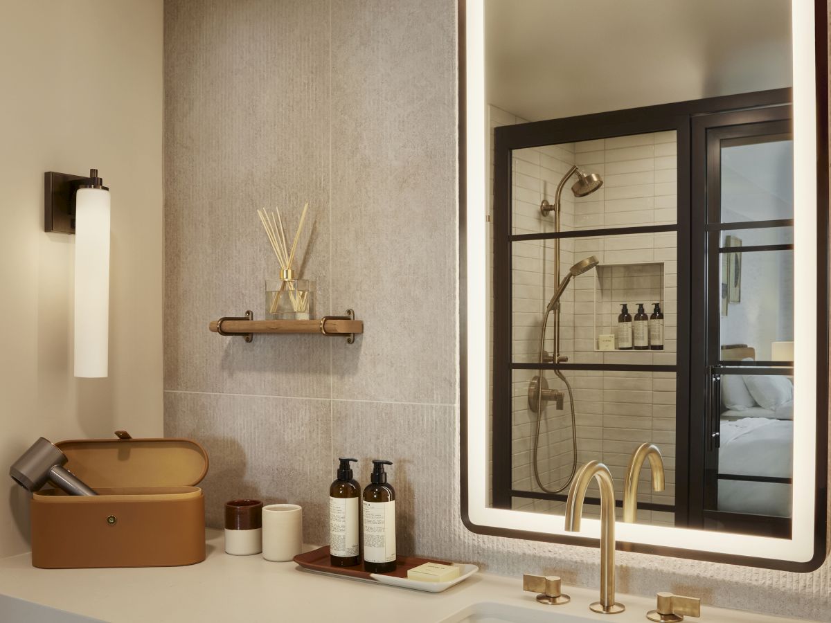 A modern bathroom features a lit mirror, stylish sink, toiletries on the counter, a suitcase-like box, and a shower visible through glass.