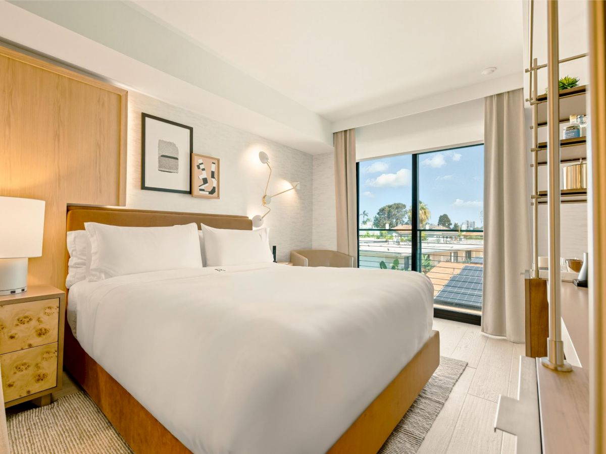 A bright hotel room with a big white bed, wooden headboard, nightstands, wall art, a small desk, and floor-to-ceiling windows with a balcony view.