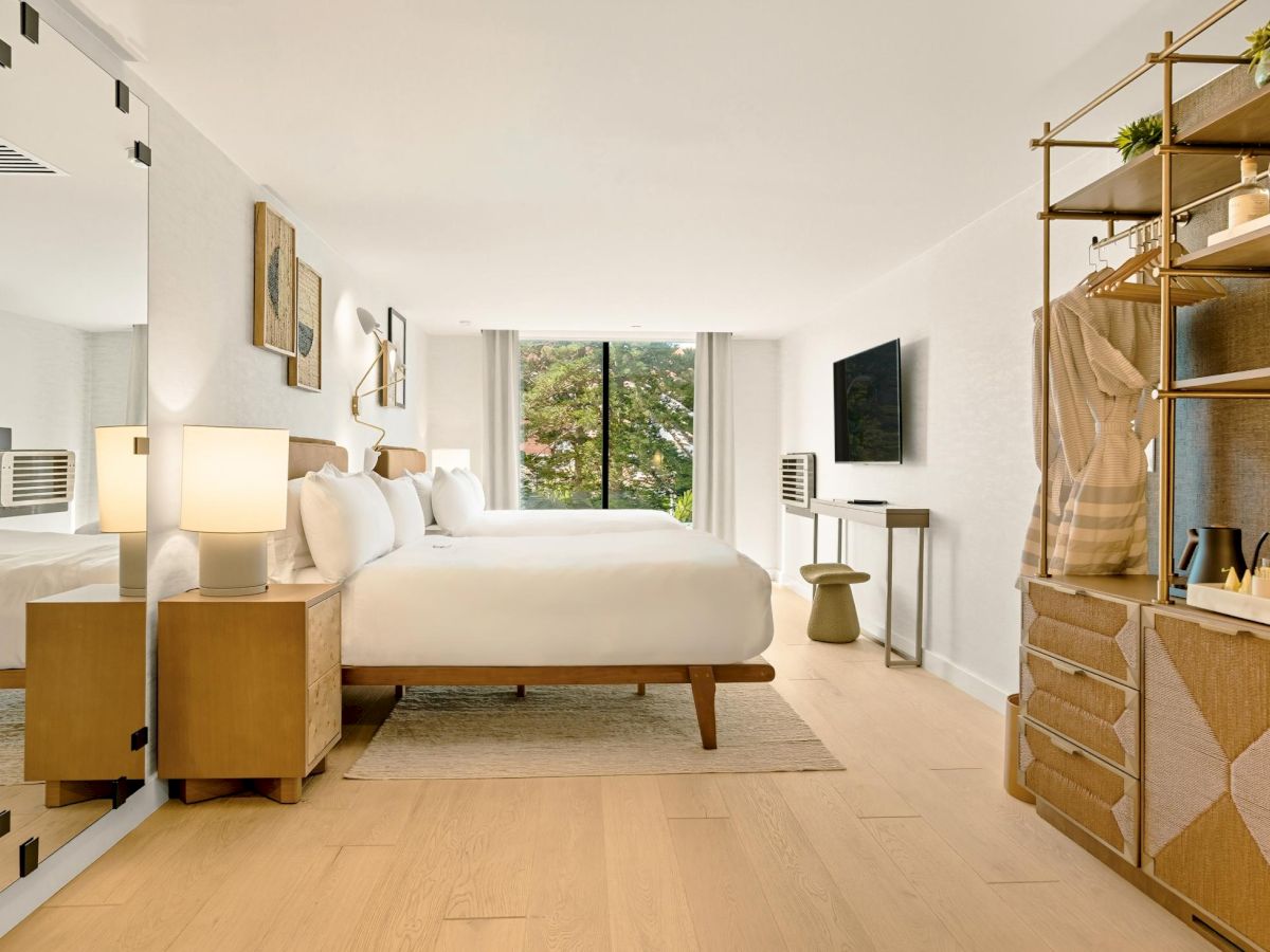 A bright, modern hotel-style bedroom with a large bed, mirrored closet, wooden floors, a desk, wall-mounted TV, and open shelving, all softly lit.