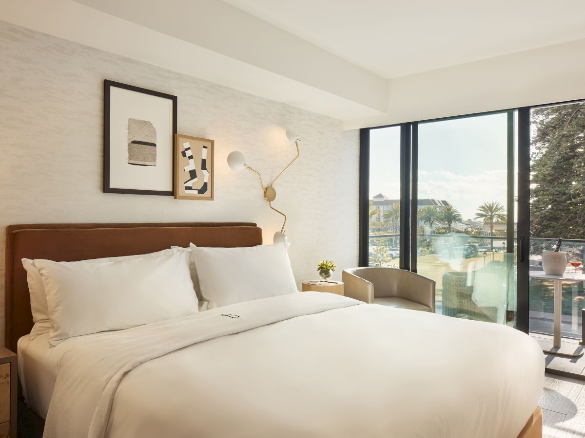 A cozy modern bedroom with a large bed, art on walls, a lamp, chair, and a view of a balcony with trees outside.