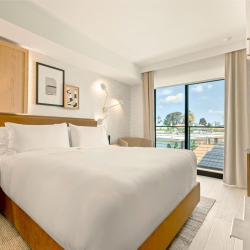 A bright hotel room with a king bed, bedside lamps, a desk, seating area, and a large window with natural light.