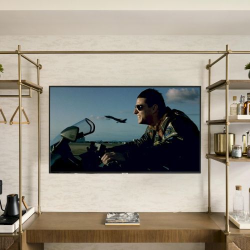 A bright modern living room with a large TV mounted on the wall showing a beach scene, flanked by two open closets and plants, windows and seating.
