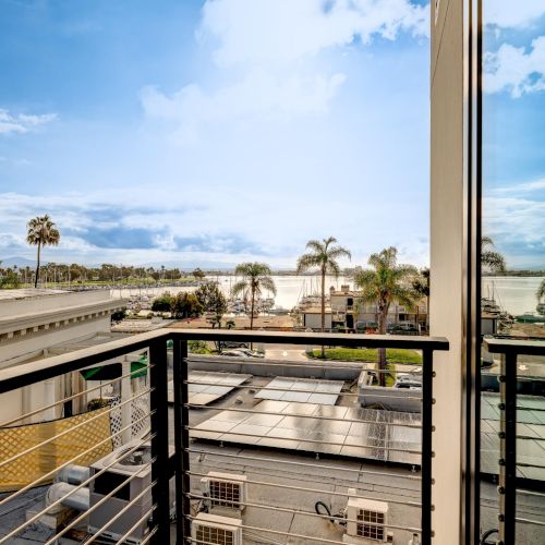 A sunny balcony view overlooking rooftops and a blue coastal sky, with a wooden railing and distant horizon.