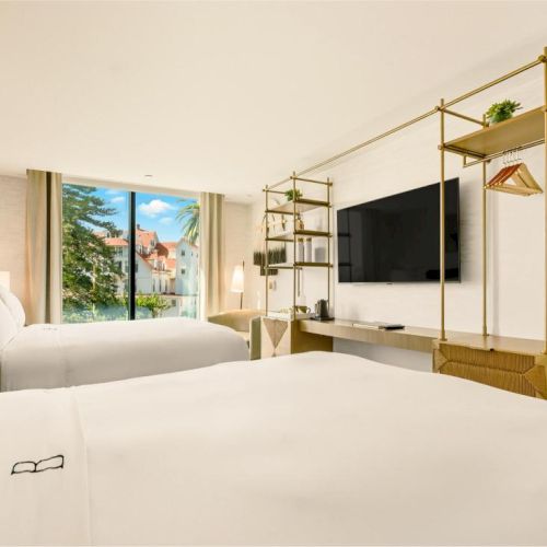 A bright hotel room with two white beds, a wall-mounted TV, open wardrobe racks, a desk, and a window showing trees and a building outside.