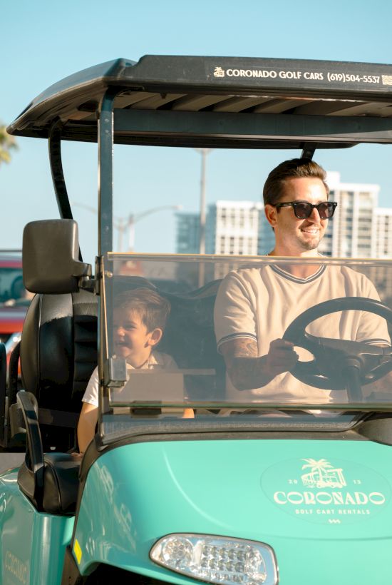 Two men ride in a turquoise golf cart with Colorado branding; the driver wears sunglasses, smiling, as they pace by sunny buildings.