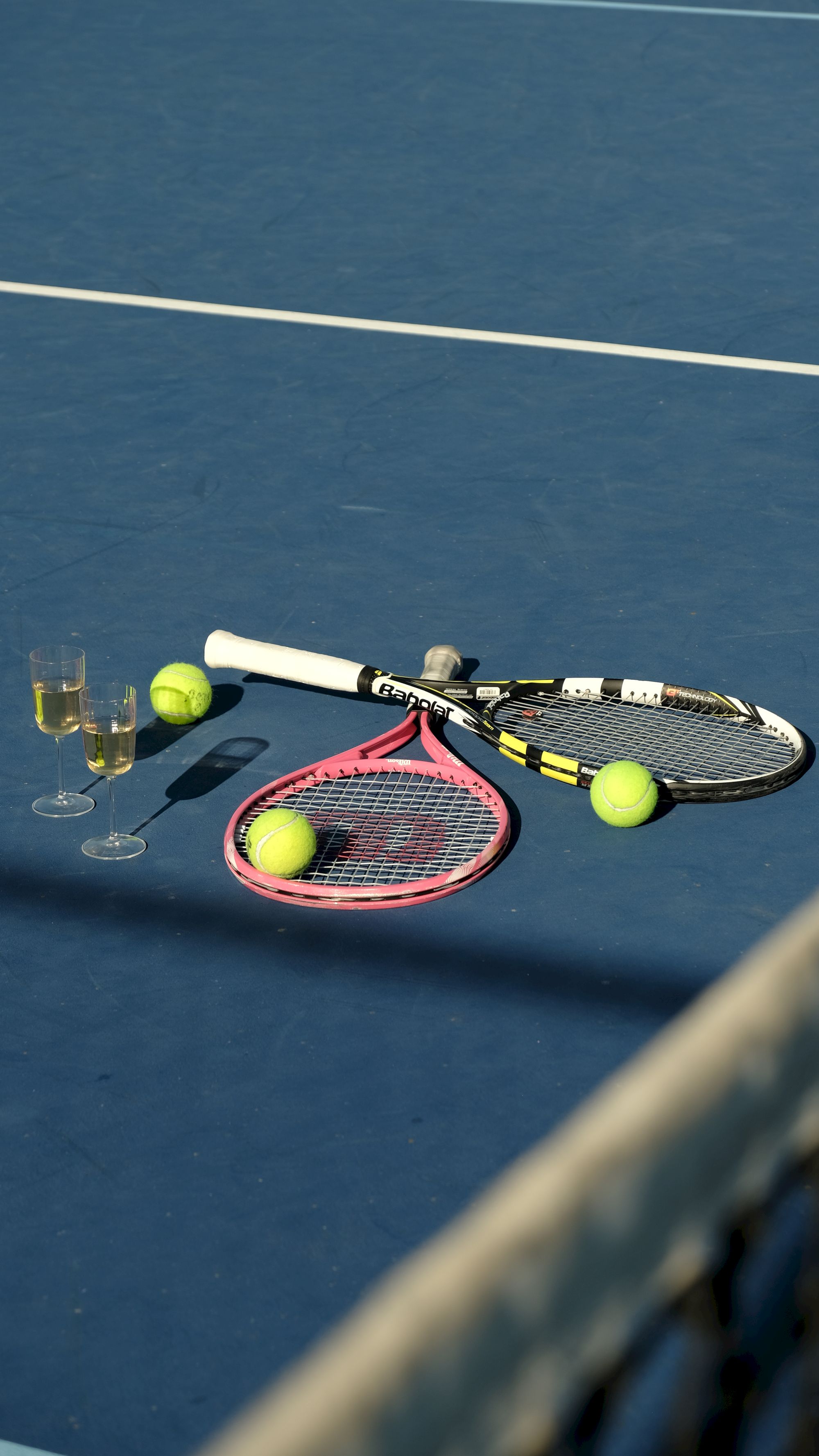 Tennis gear lies on a blue court: racquets, yellow tennis balls, a glow of sunlight, and two glasses of champagne resting near the lines.