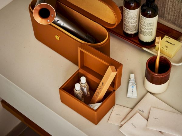 A leather vanity case, open with grooming items like bottles, a tube, and small boxes; a matching brown makeup box and tray sit on a counter.