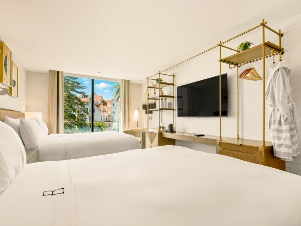 A bright hotel room with two white beds, a wall-mounted TV, open wardrobe racks, a window showing trees outside, and a robe hanging by the door.