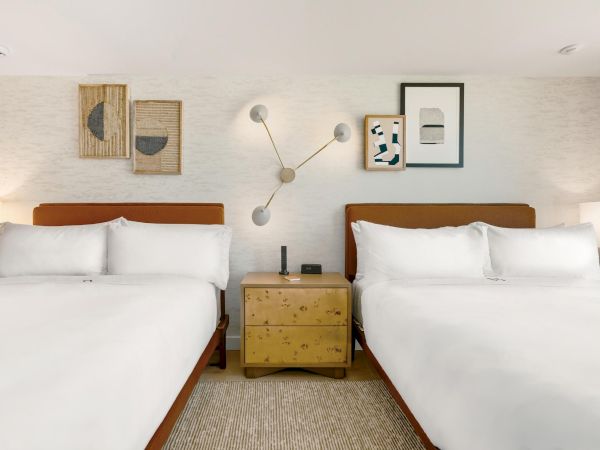 Two neatly made side-by-side hotel beds, a wooden nightstand between them, wall art, lamps, and a modern sconce on a light textured wall.