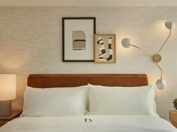 A neatly made bed with white linens, a brown headboard, two pillows, nightstands, wall art above, and a modern wall lamp in a calm bedroom.