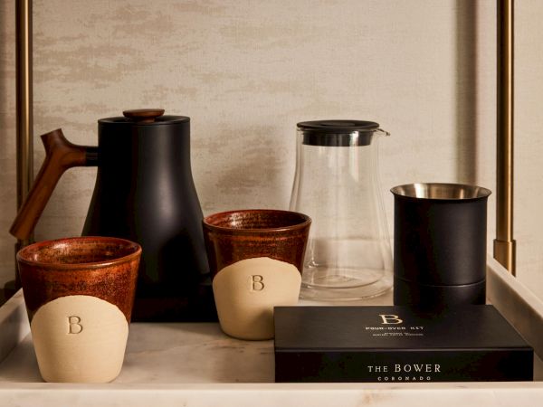 A stylish coffee station with two ceramic cups, a black kettle, a glass carafe, a white-tipped cup, and a black box labeled &ldquo;The Bower&rdquo; on a tray.