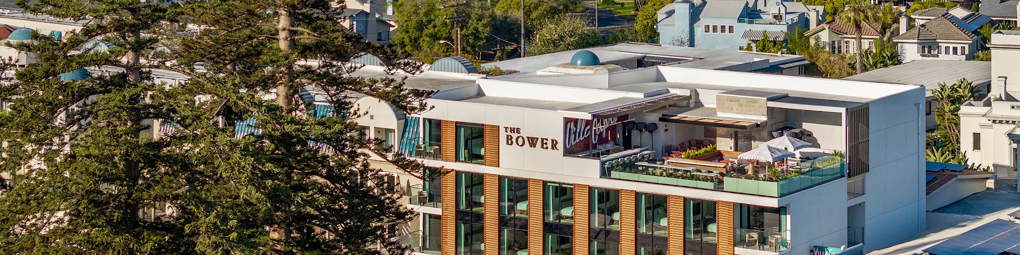 A modern hotel with a glassy facade sits in a coastal suburb, trees frame the foreground, and a city skyline rises in the distance.
