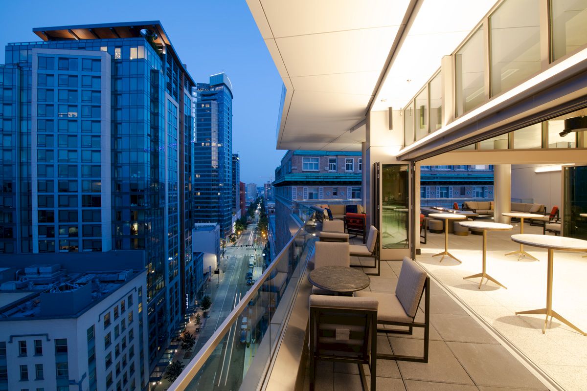 A modern outdoor balcony with tables and chairs overlooks a city street lined with tall buildings at twilight.