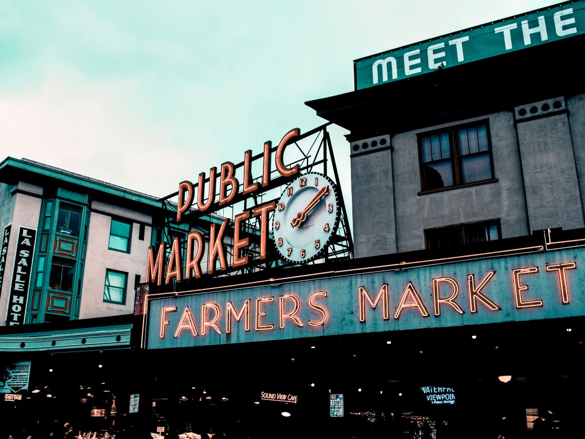 The image shows a historic public market with neon signs for "Public Market" and "Farmers Market" and a clock on the facade of a building.