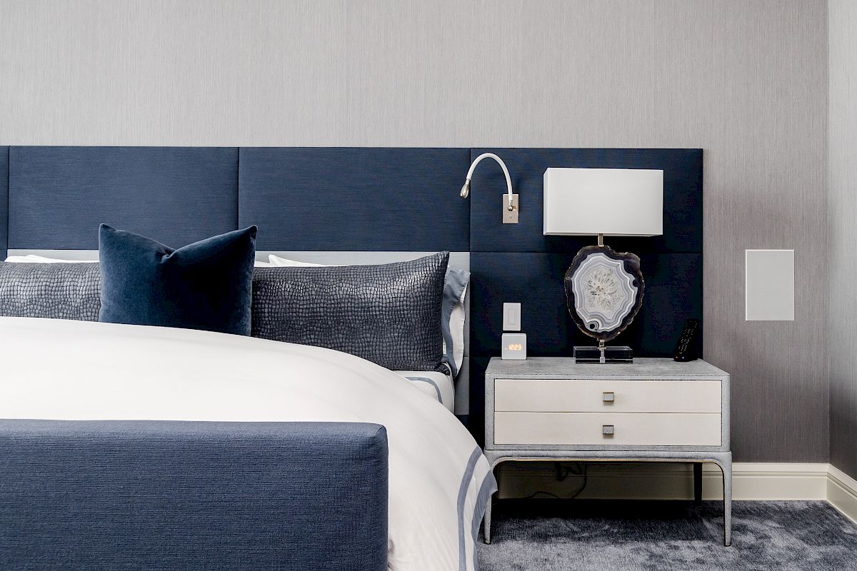 A modern bedroom with a navy bed, gray walls, and a white nightstand featuring a lamp and decorative item on a plush carpet.