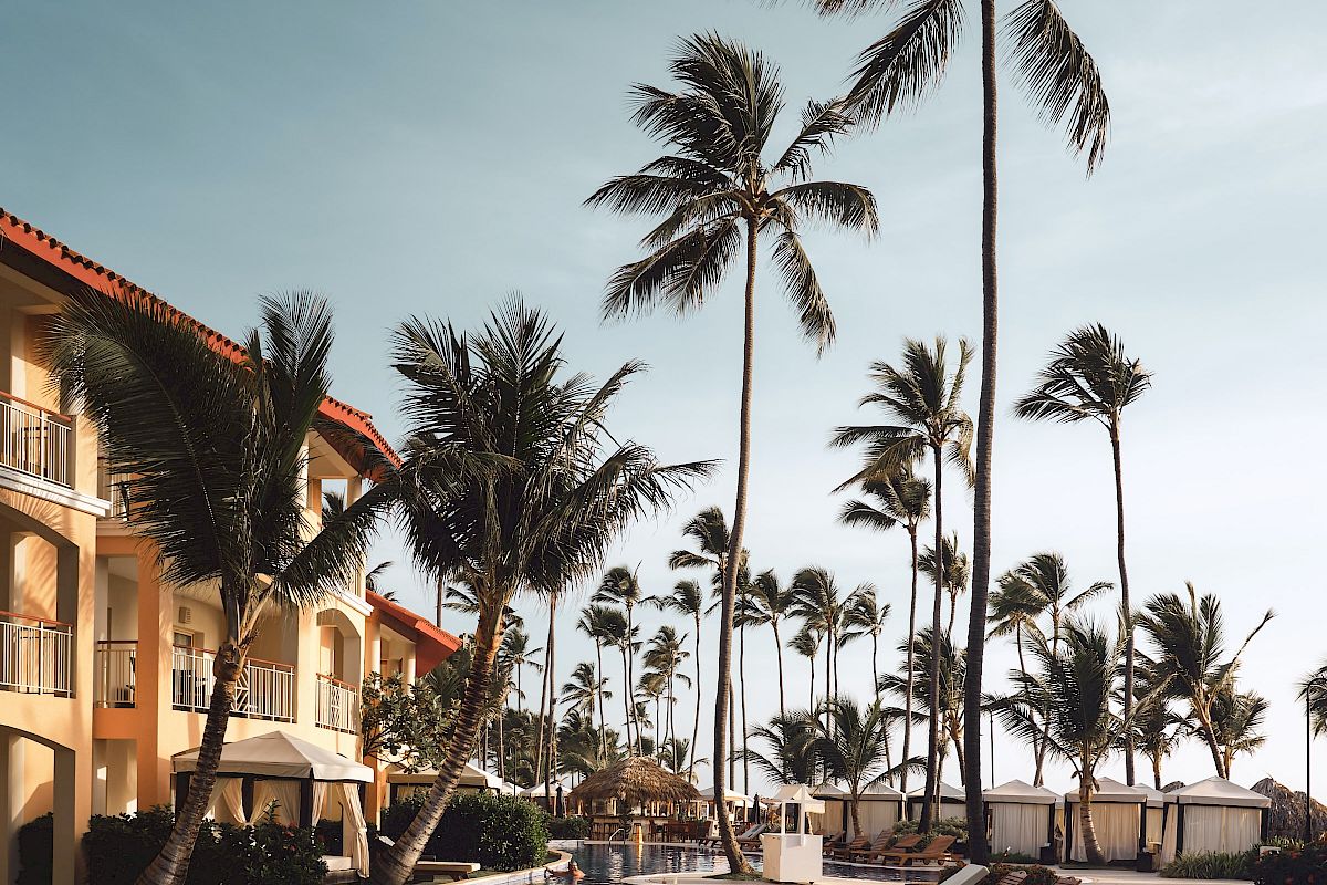A resort pool surrounded by palm trees and lounge chairs under a clear sky, next to a building with balconies.